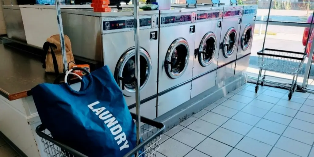 Tas laundry biru di troli, di depan deretan mesin cuci.